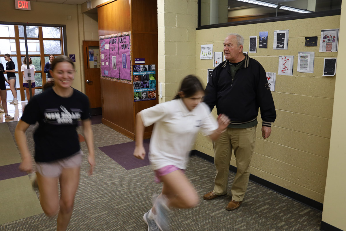 Coach Clem Papineau (in the back right) wears a black jacket and khaki pants as two high school girls, one in a black shirt and one in a white shirt, run past him.