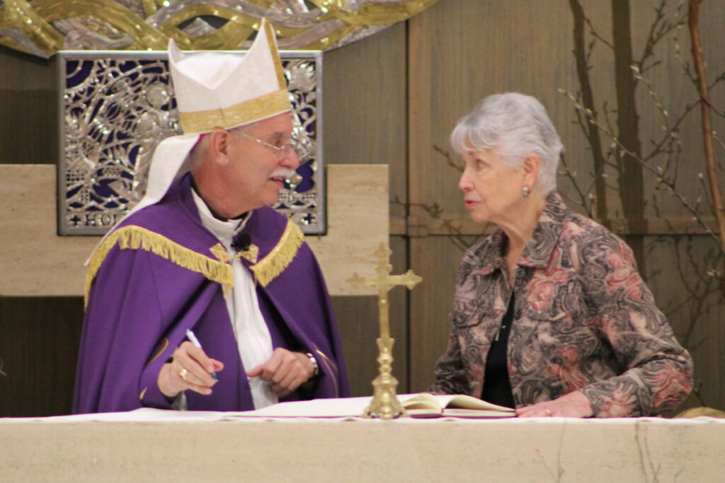 Gail Zanoff, an older white woman in a floral shirt, stands behind the altar with Bishop Anthony B. Taylor, who is wearing a purple clerical robe.