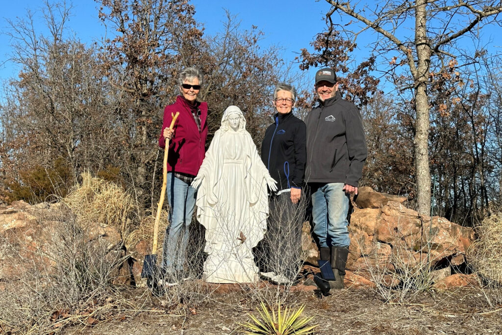 Three older Catholics hold shovels and outdoor equipment as they stand beside a statue of the Blessed Virgin Mary on retreat center grounds