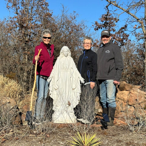 Three older Catholics hold shovels and outdoor equipment as they stand beside a statue of the Blessed Virgin Mary on retreat center grounds