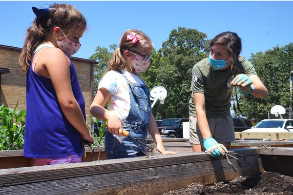 Rising fourth grade students Clara Hum (left) and Blair Bramlett, both 9, listen Aug. 6 to AmeriCorps volunteer Carey McKay explain that old roots can be added to the compost pile at the Immaculate Conception School garden in North Little Rock.