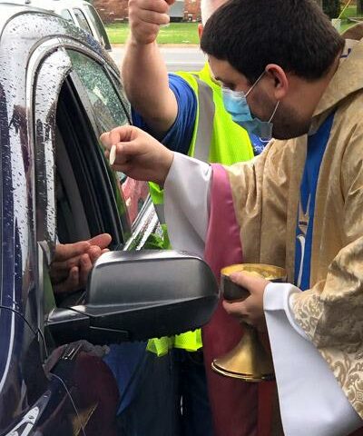 Father Alejandro Puello, pastor of St. Anne Church in North Little Rock, distributes Communion May 17 during a drive-in Mass he celebrated in the rainy church parking lot. Bill Ortner holds an umbrella to protect his pastor.