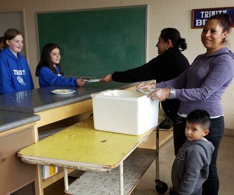 Trinity seventh-graders Ava Edwards (left) and Karlee Rusin get pupusas for lunch at Trinity Catholic School, prepared by volunteers Agueda Guerrero and Angie Manjarrez, accompanied by 3-year-old Andrew Manjarrez, April 10.