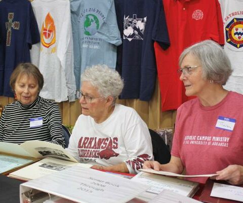 Anne-Marie Candido, Mary Adams and Theresa Cronan look through historical documents from St. Thomas Aquinas University Parish in Fayetteville Oct. 21. T-shirts that have been made through the years for the students hung in display behind them.